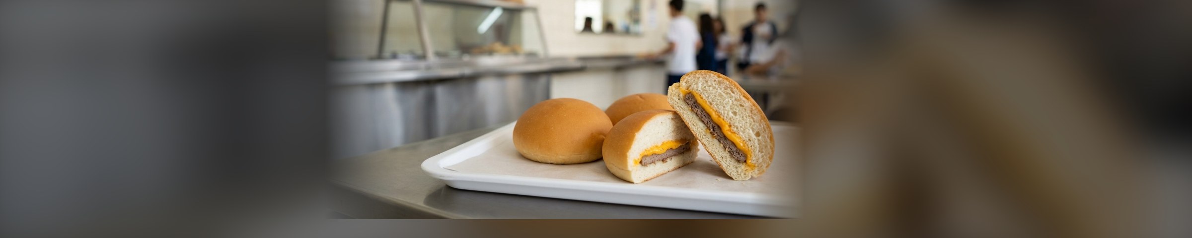 Hamburgão de cheddar partido ao meio em bandeja, com o refeitório de uma cantina escolar desfocado ao fundo
