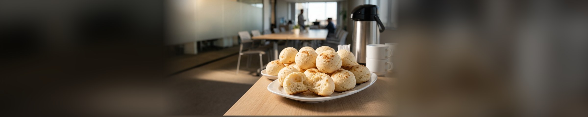 Travessa de pães de queijo, um deles partido mostrando o miolo, numa mesa de escritório preparada para a confraternização